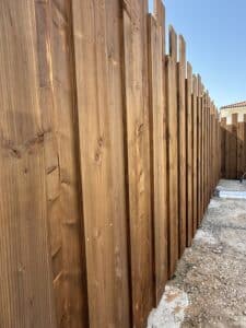 Clôture de jardin en bois marron clair, panneaux verticaux à hauteur variable sous un ciel bleu dégagé. Sol de gravier au premier plan et toiture de maison au loin.