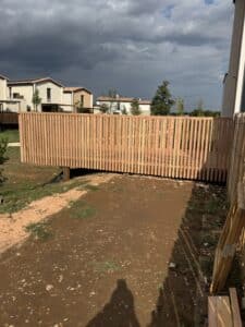 Clôture en bois clair devant des maisons modernes. Sol terreux, herbe clairsemée et ciel nuageux contrasté, avec zones sombres.