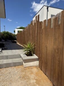 Clôture en bois foncé protégeant une terrasse moderne grise avec un yucca en pot et un ballon de foot sous un ciel bleu ensoleillé.