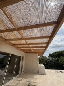 Pergola bois avec toit en canisses filtrant le soleil sur une terrasse carrelée. Baie vitrée, jardin et ciel nuageux.