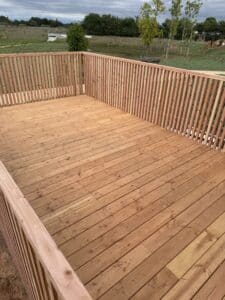 Terrasse en bois clair neuve avec balustrade à lattes verticales. Vue sur un paysage verdoyant, arbres et ciel nuageux.