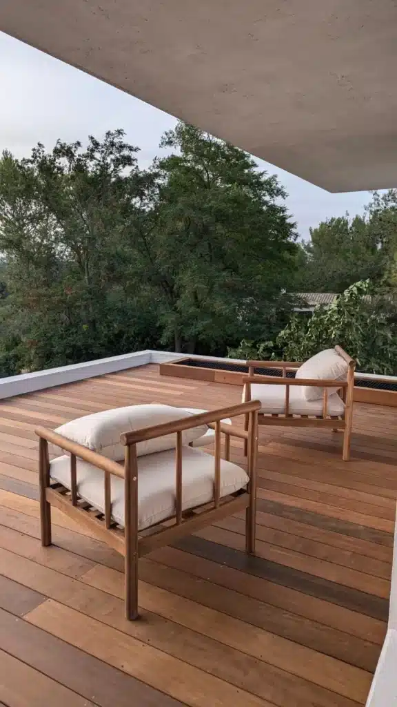 Terrasse moderne en bois avec deux chaises longues et coussins crème, entourée d'arbres luxuriants sous un ciel couvert.