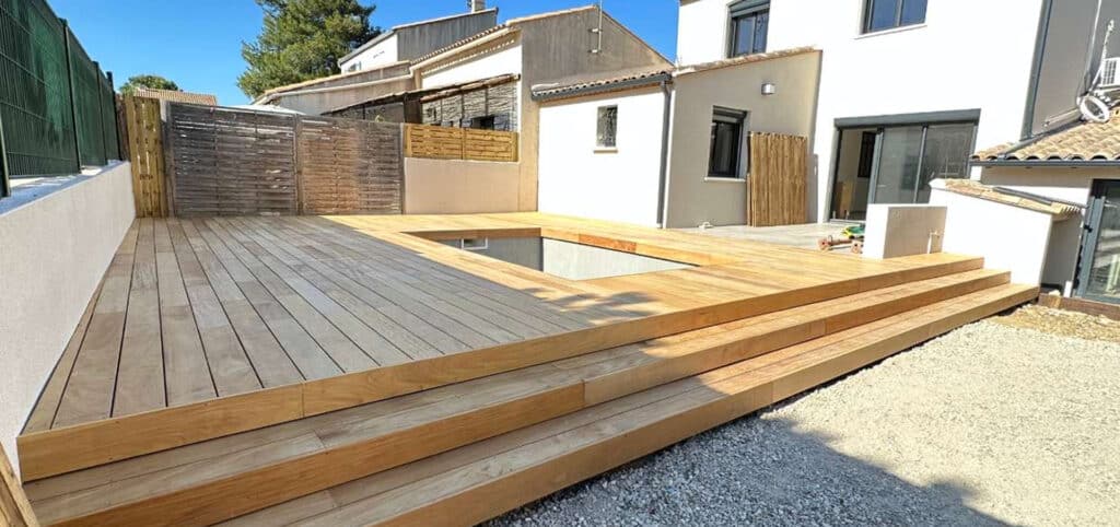 Grande terrasse en bois clair avec un bassin intégré et plusieurs marches menant à un jardin gravillonné, devant une maison moderne.