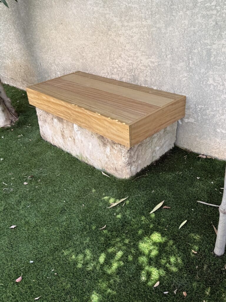 Banc de jardin en bois clair sur base de pierre, devant un mur texturé, avec pelouse synthétique verte et feuilles au sol.