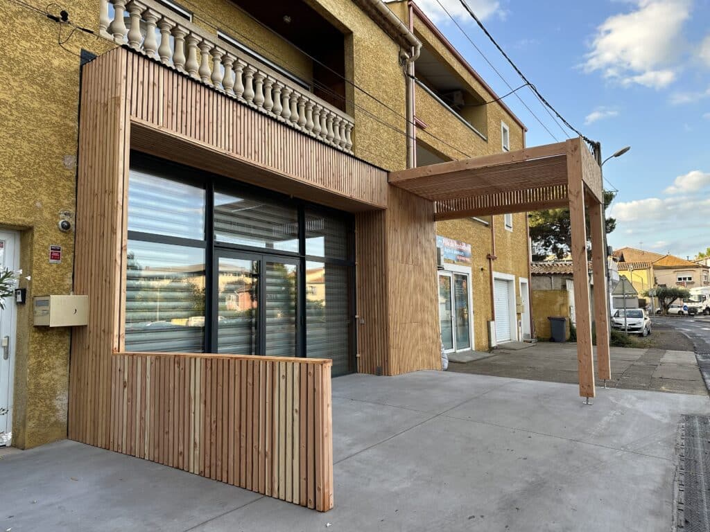 Façade rénovée à Agde : bois, vitrines et pôle productivité Façade jaune rénovée avec bardage bois, grandes vitrines et pergola. Balcon balustrade béton, panneau "Pôle de Productivité Agde & alentours".