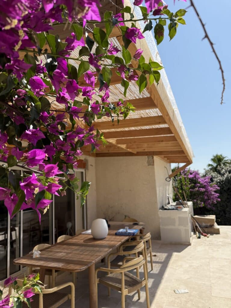 Bougainvillier violet vif encadrant une terrasse ensoleillée. Table et chaises en bois sous une pergola. Ambiance extérieure lumineuse.