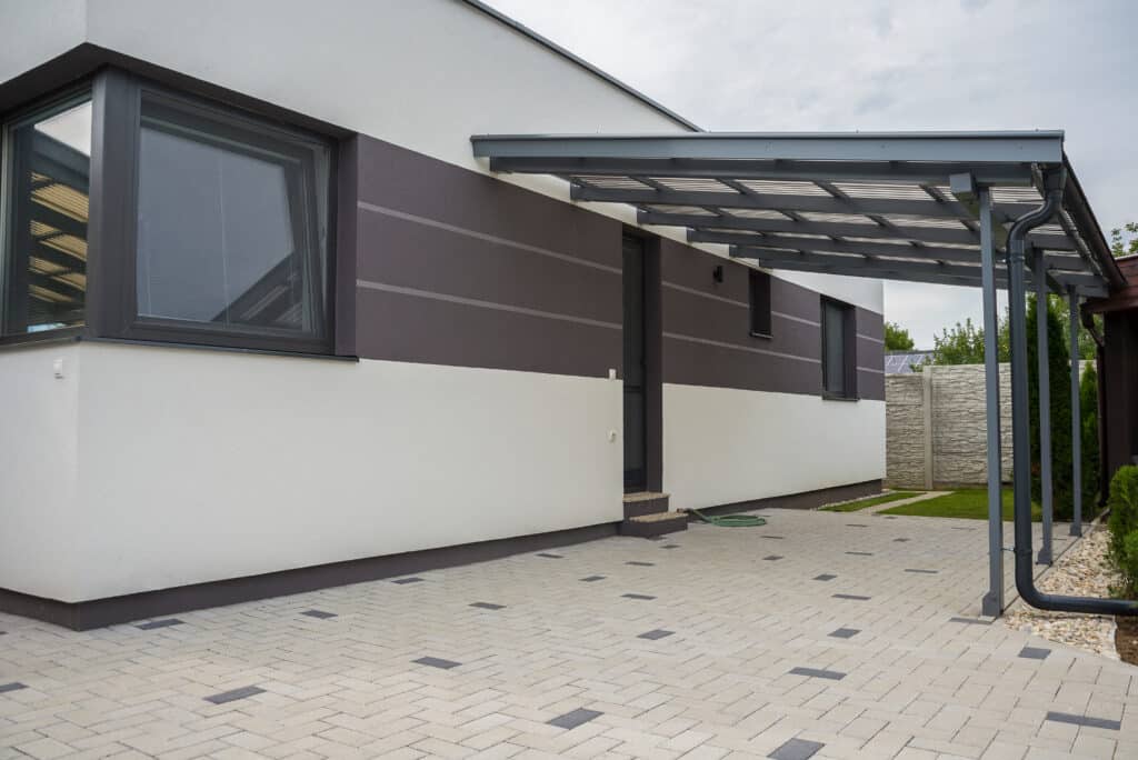 Façade moderne d'une maison blanche et marron avec fenêtres sombres, sous un carport gris. Allée pavée clair avec motifs foncés, jardin.