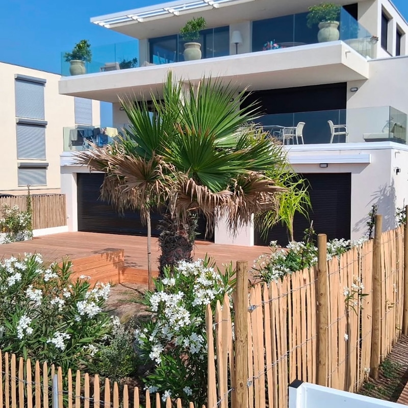 Maison moderne blanche avec grandes baies vitrées et terrasses. Jardin méditerranéen avec palmiers, fleurs blanches et clôture en bois. Terrasse au sol en bois.