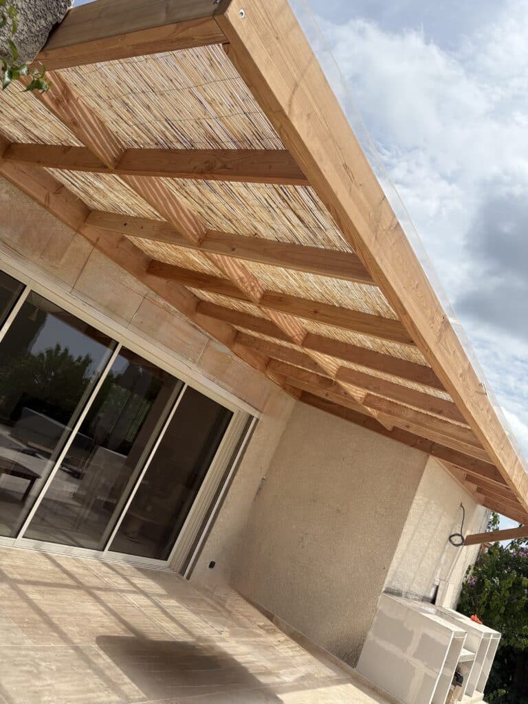 Pergola moderne en bois et bambou au-dessus d'une terrasse carrelée. Toiture transparente, grandes portes coulissantes reflétant le jardin.