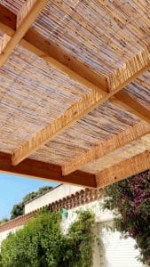 Pergola en bois avec canisse ajourée, diffusant la lumière du soleil sur une façade méditerranéenne ornée d'un nichoir et de bougainvilliers.