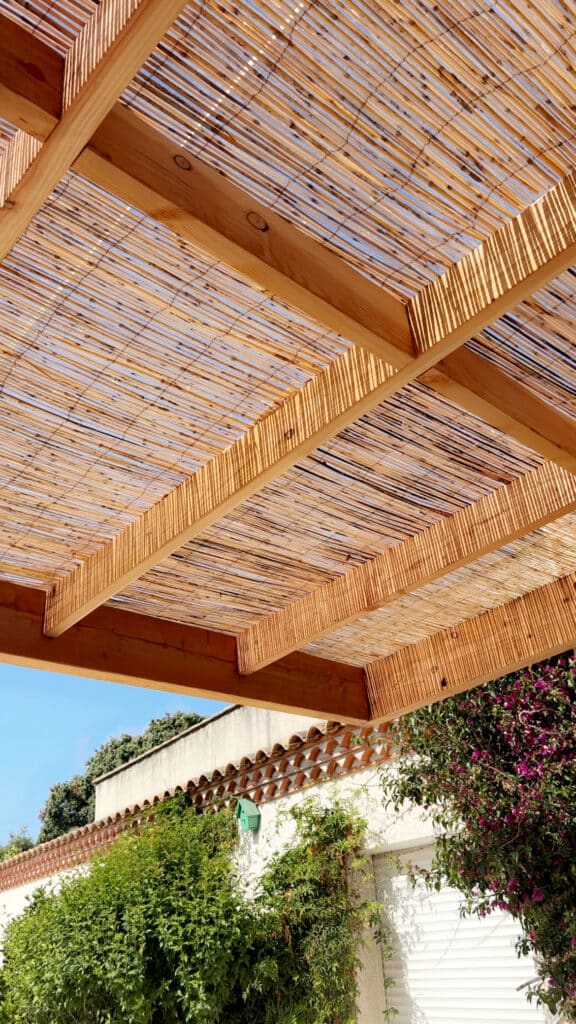 Pergola en bois avec canisse ajourée, diffusant la lumière du soleil sur une façade méditerranéenne ornée d'un nichoir et de bougainvilliers.