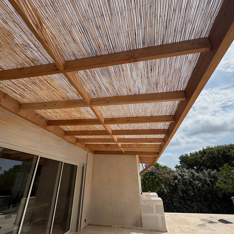 Pergola moderne en bois clair avec toit en canisses, ombrageant une terrasse avec grande baie vitrée et verdure luxuriante.