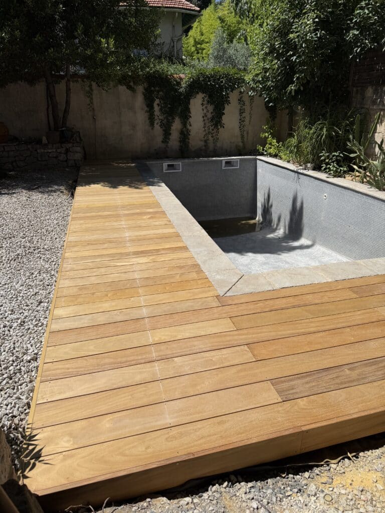 Piscine vide carrelée de mosaïque grise avec nouvelle terrasse en bois clair. Gravier blanc, murs végétalisés et arbres ensoleillés.