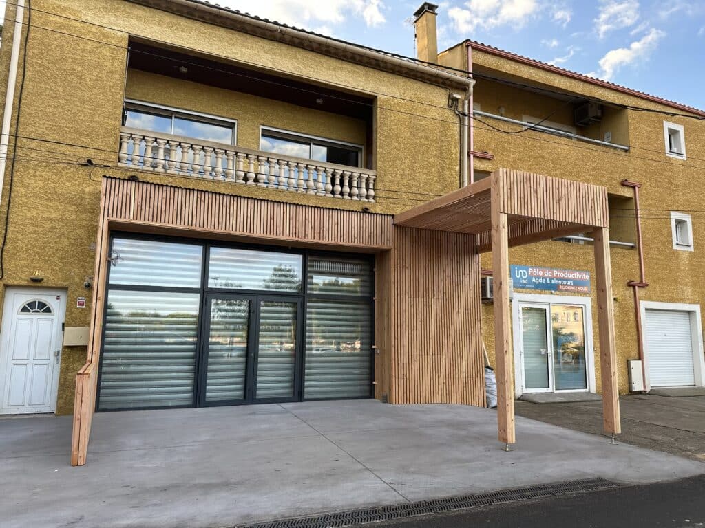 Façade jaune bâtiment commercial, structure bois moderne, vitrines, balcon. Signe: Pôle de Productivité Agde & alentours, REJOIGNEZ NOUS.