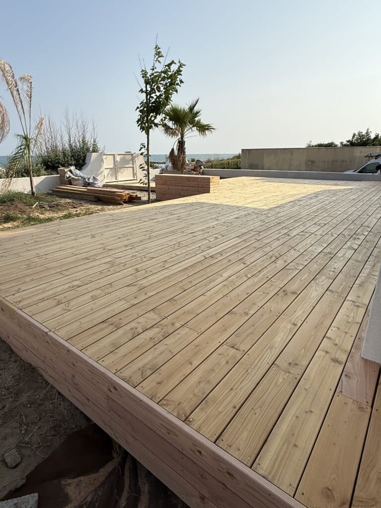 Nouvelle terrasse en bois clair au bord de mer sous un ciel bleu dégagé. Aménagement extérieur avec plantes et vue sur l'océan.