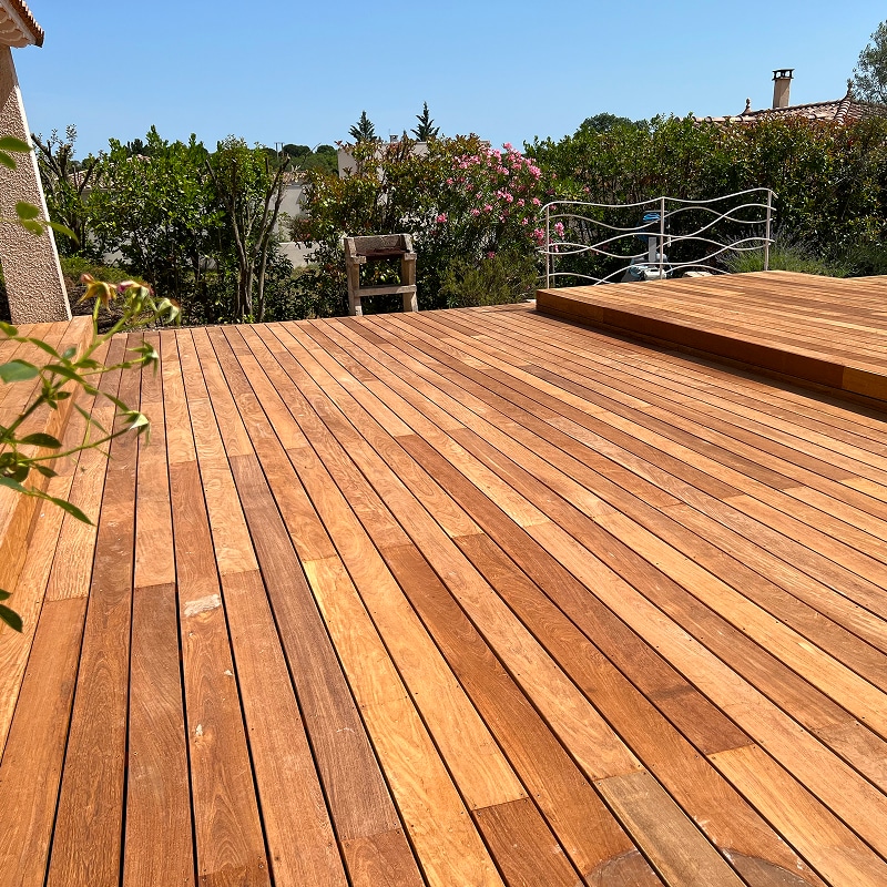 Spacieuse terrasse en bois clair sous le soleil, avec jardin luxuriant, barbecue en pierre et garde-corps blanc ondulé.